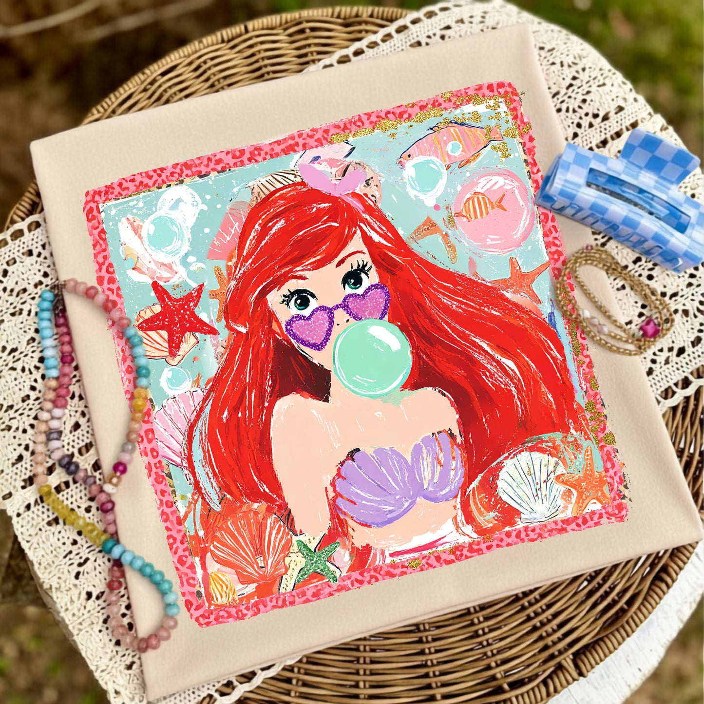 a graphic tshirt laying in a basket. the image on the tshirt is a colorful, handdrawn, cartoon image of a redhaired mermaid blowing a bubble.