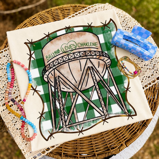Folded ivory graphic T-shirt featuring a vintage-style water tower with the phrase "BillyBob Loves Charlene" against a green gingham background, styled on a wicker tray with colorful beaded bracelets and coastal-inspired accessories.