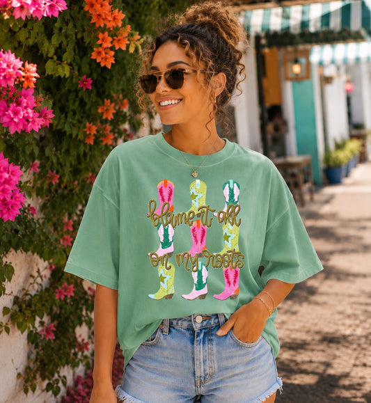 Woman standing in front of a floral wall wearing a green tshirt with colorful boots and the words blame it all on my roots on it.