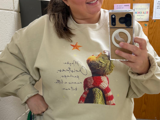 Person wearing a beige sweatshirt with a graphic of a the Grinch wearing a red scarf.
