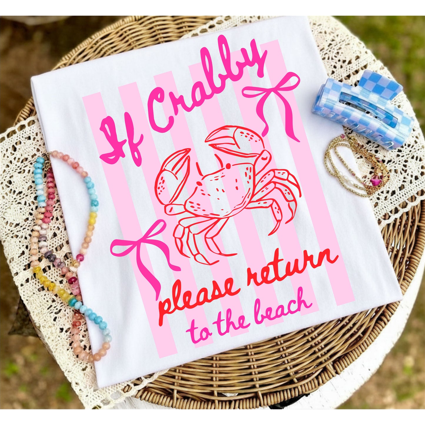 Folded white graphic T-shirt with pink "If Crabby, Please Return to the Beach" crab design displayed on a woven tray with colorful beaded jewelry and beach-inspired accessories.