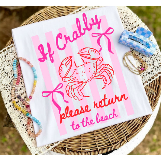 Folded white graphic T-shirt with pink "If Crabby, Please Return to the Beach" crab design displayed on a woven tray with colorful beaded jewelry and beach-inspired accessories.