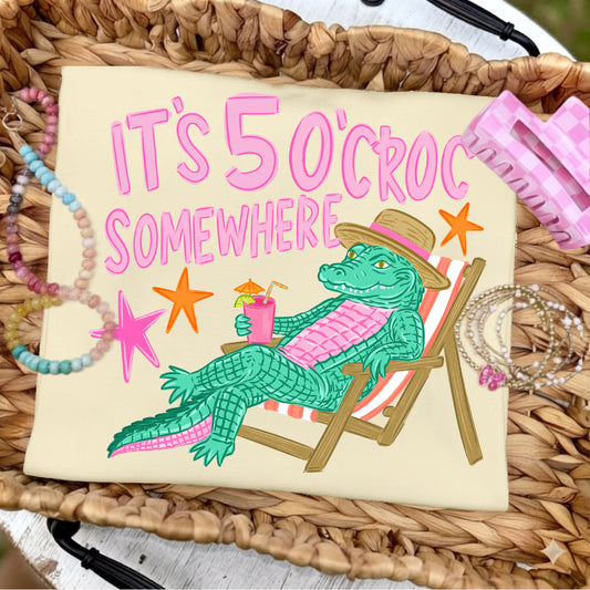 Folded butter yellow graphic T-shirt featuring the colorful "It's 5 O'Croc Somewhere" design with a lounging alligator in a beach chair, styled on a woven tray with pastel beaded bracelets, jewelry, and pink beach-inspired accessories.