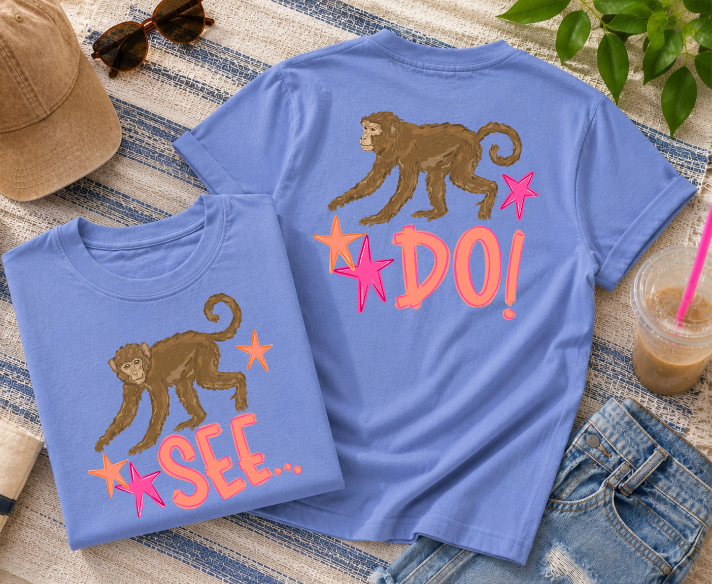 Folded and back-view blue graphic T-shirts displaying the playful "Monkey See, Monkey Do" front-and-back monkey design, styled with a neutral hat, sunglasses, iced coffee, denim shorts, and coastal-inspired accessories.