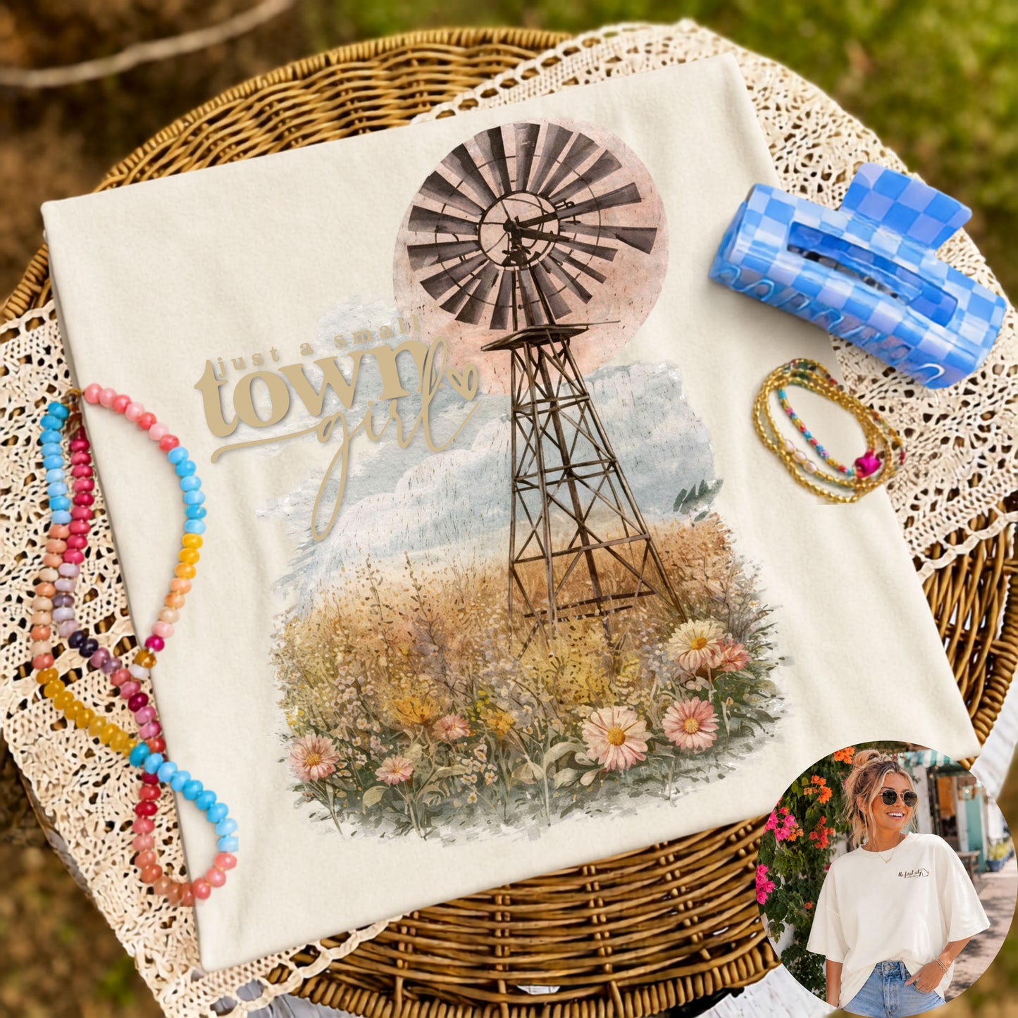Flat lay of a cream graphic t-shirt featuring a vintage windmill surrounded by watercolor wildflowers with the phrase "Just a Small Town Girl," styled with colorful beaded bracelets, gold bracelets, and a blue gingham hair clip on a wicker table with lace fabric.