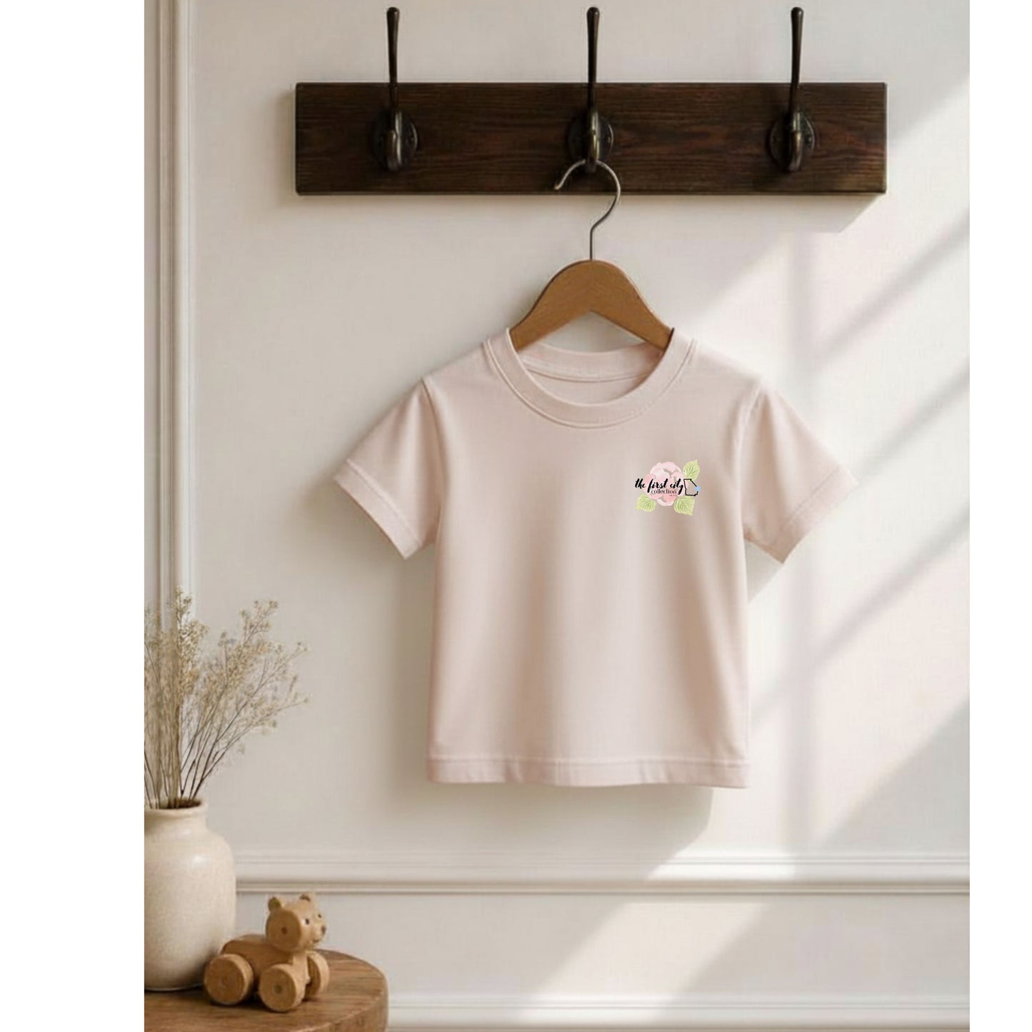 Beige t-shirt with a small logo on a wooden hanger against a white wall.
