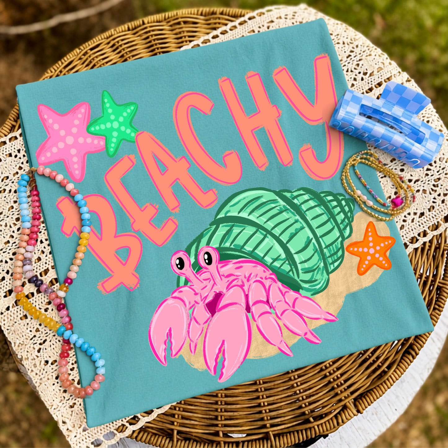 Folded teal graphic T-shirt featuring a colorful pink hermit crab inside a mint green seashell with the word "Beachy," styled on a wicker tray with colorful beaded bracelets, coastal accessories, and lace fabric.