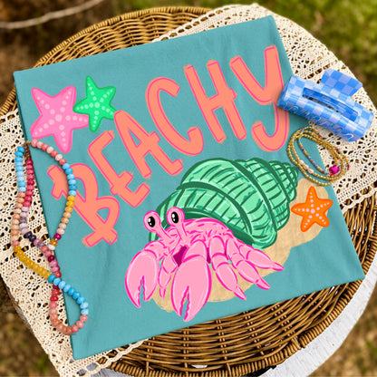 Folded teal graphic T-shirt featuring a colorful pink hermit crab inside a mint green seashell with the word "Beachy," styled on a wicker tray with colorful beaded bracelets, coastal accessories, and lace fabric.