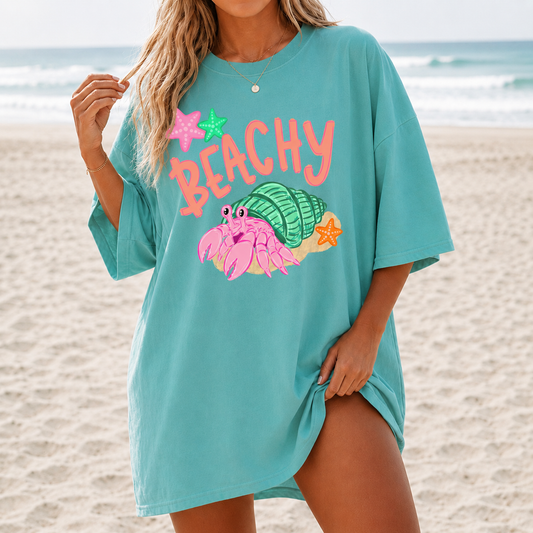 Woman standing on a beach wearing a teal t-shirt with a hermit crab and the word beachy on it.