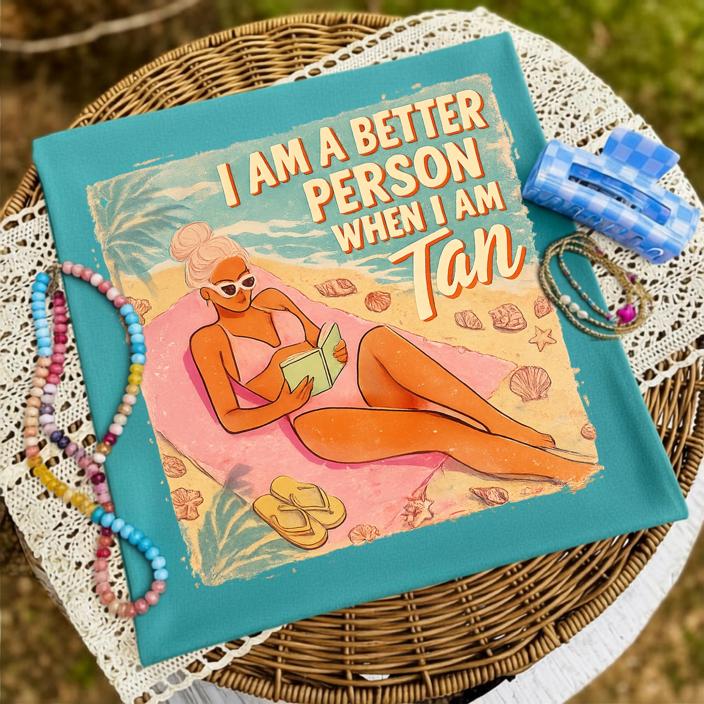 Folded teal graphic T-shirt featuring an illustrated woman relaxing on a beach towel reading a book with the phrase "I Am a Better Person When I Am Tan," styled on a wicker tray with colorful beaded bracelets and coastal-inspired accessories.