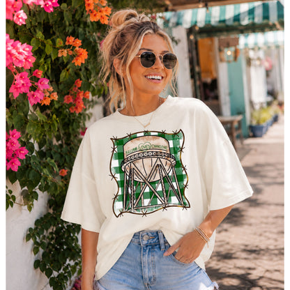 a woman standing in front of a wall with flowers on it wearing an ivory colored t-shirt with artwork showing a water tower that says Billy Bob Loves Charlene against a green and white gingham background.