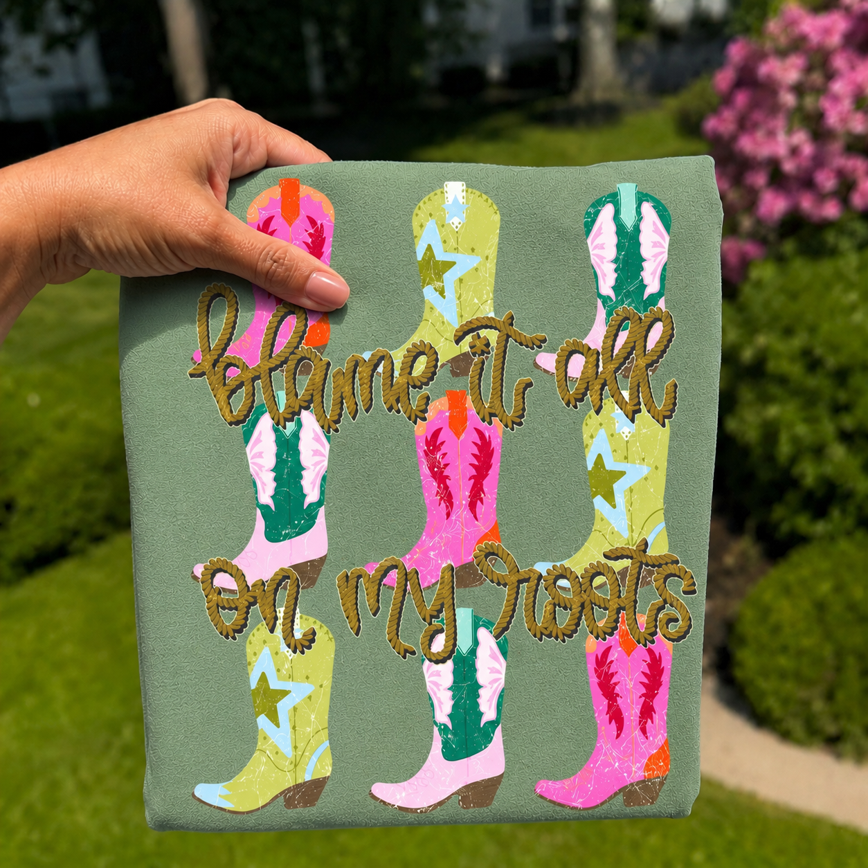 Folded sage green graphic T-shirt featuring colorful cowboy boots with rustic rope lettering that reads "Blame It On My Roots," photographed outdoors with a hand holding the shirt against a blurred garden background.