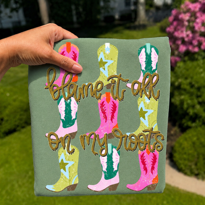 Folded sage green graphic T-shirt featuring colorful cowboy boots with rustic rope lettering that reads "Blame It On My Roots," photographed outdoors with a hand holding the shirt against a blurred garden background.