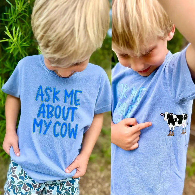 Child wearing a blue t-shirt with 'Ask Me About My Cow' text and a cow illustration.
