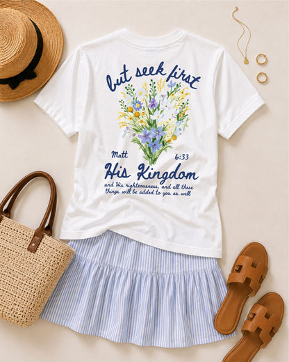 White Seek First His Kingdom Christian graphic tee featuring Matthew 6:33 and colorful wildflowers, styled with a blue striped skirt, tan sandals, woven tote, straw hat, and gold jewelry.