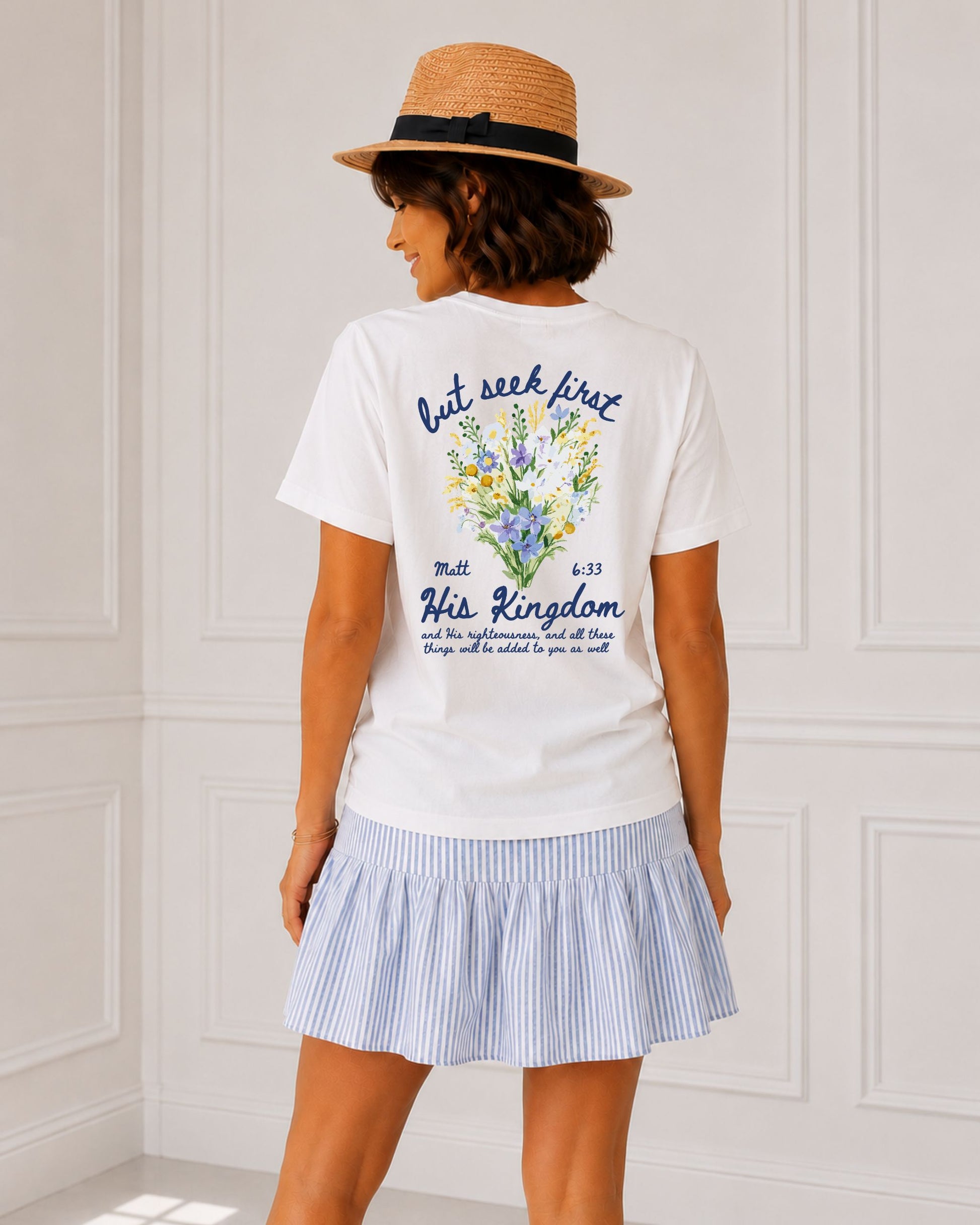 Back view of white Seek First His Kingdom Christian tee featuring Matthew 6:33 and colorful wildflowers, styled with a blue striped skirt and straw hat.