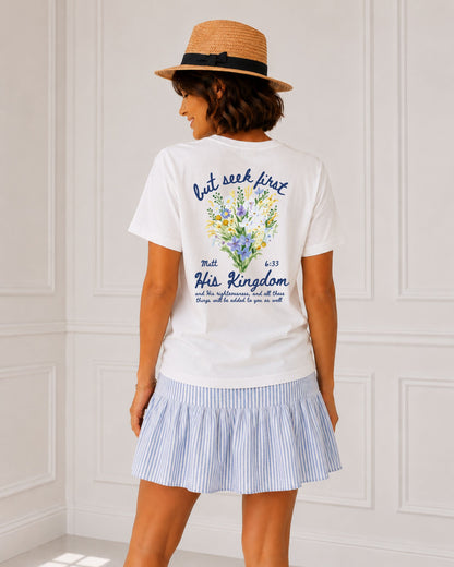 Back view of white Seek First His Kingdom Christian tee featuring Matthew 6:33 and colorful wildflowers, styled with a blue striped skirt and straw hat.
