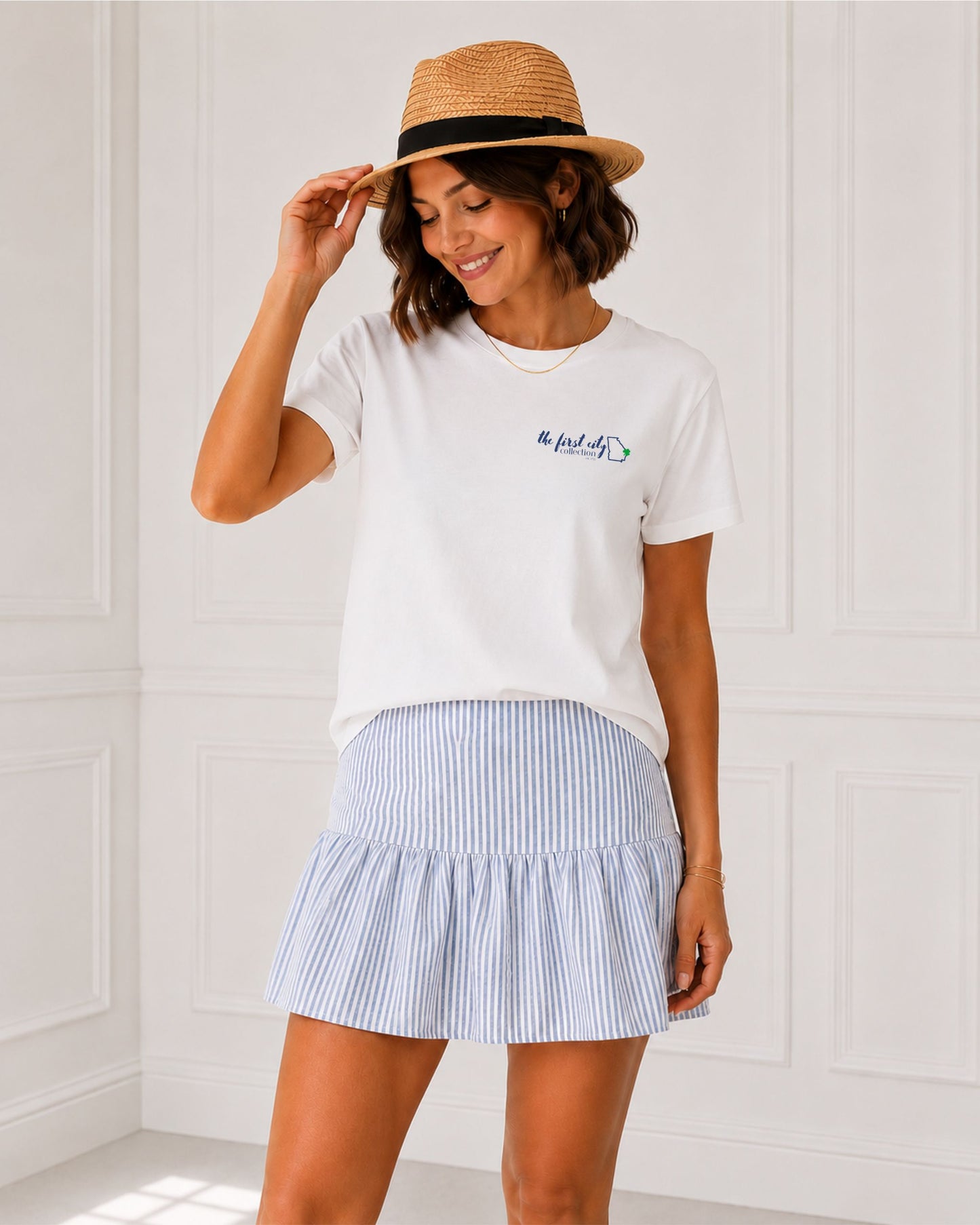 Woman modeling the white Seek First His Kingdom Christian tee with The First City Collection chest logo, styled with a blue striped skirt and straw hat.