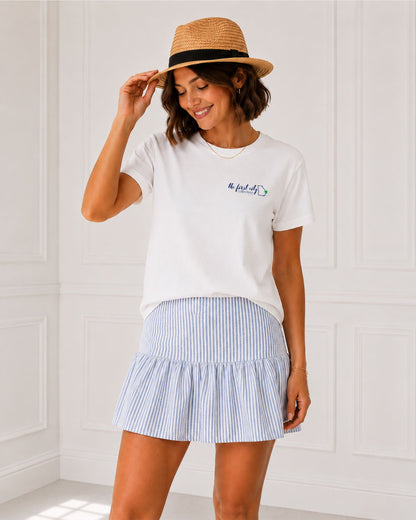 Woman modeling the white Seek First His Kingdom Christian tee with The First City Collection chest logo, styled with a blue striped skirt and straw hat.
