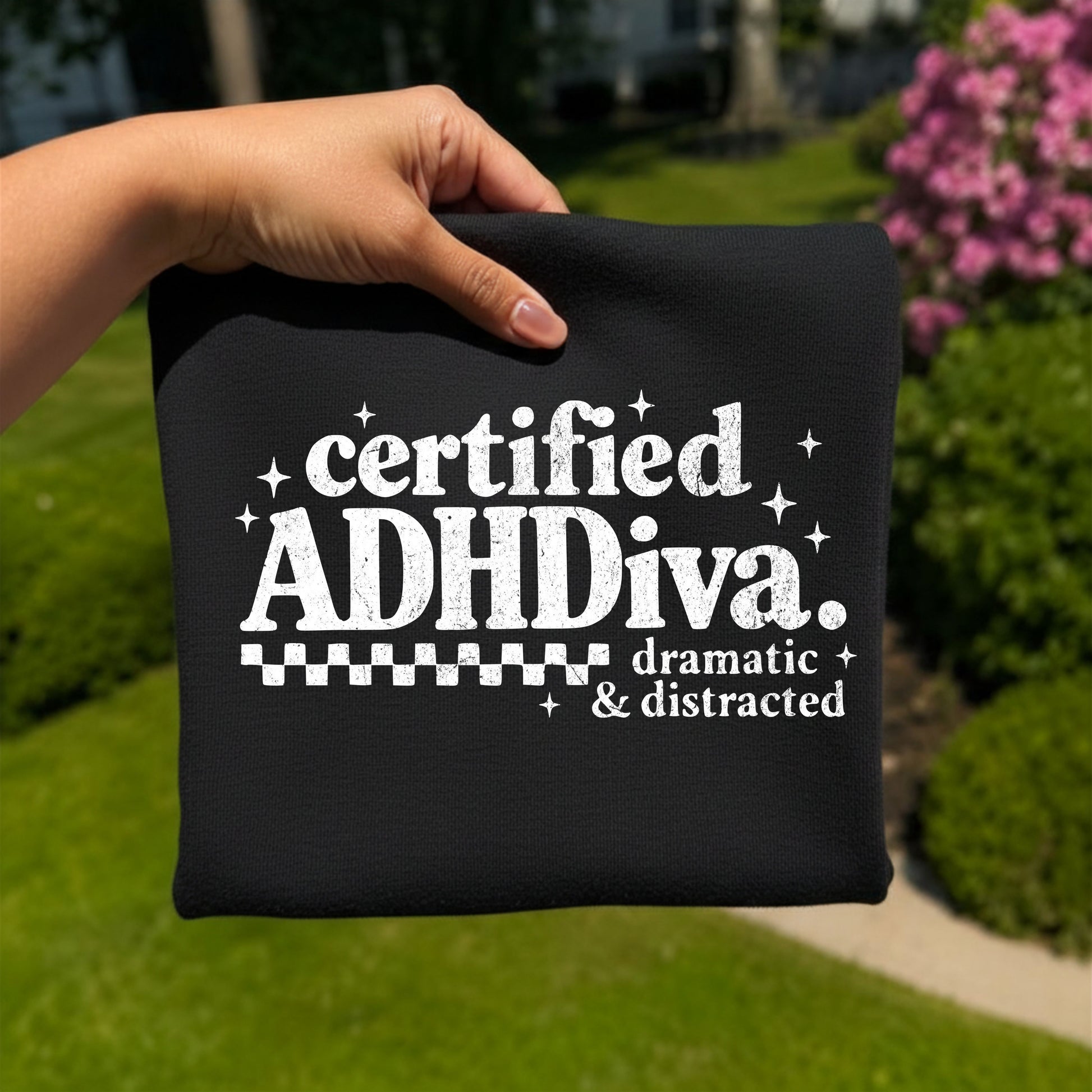 Folded black graphic T-shirt featuring distressed white typography that reads "Certified ADHDiva. Dramatic & Distracted," photographed outdoors with a hand holding the shirt against a blurred garden background.