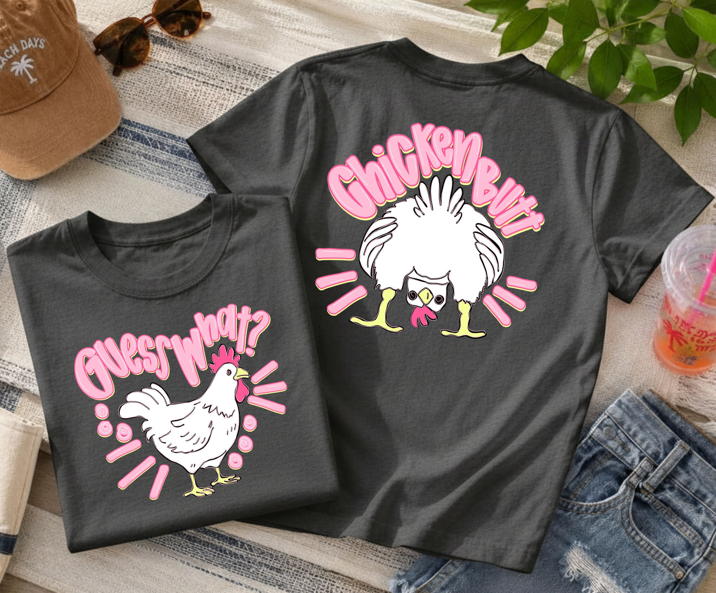 Charcoal gray front-and-back graphic T-shirt featuring a white chicken on the front with the phrase "Guess What?" and a fluffy chicken from the back with the phrase "Chicken Butt," displayed with denim shorts, sunglasses, a beach hat, and a summer drink.

