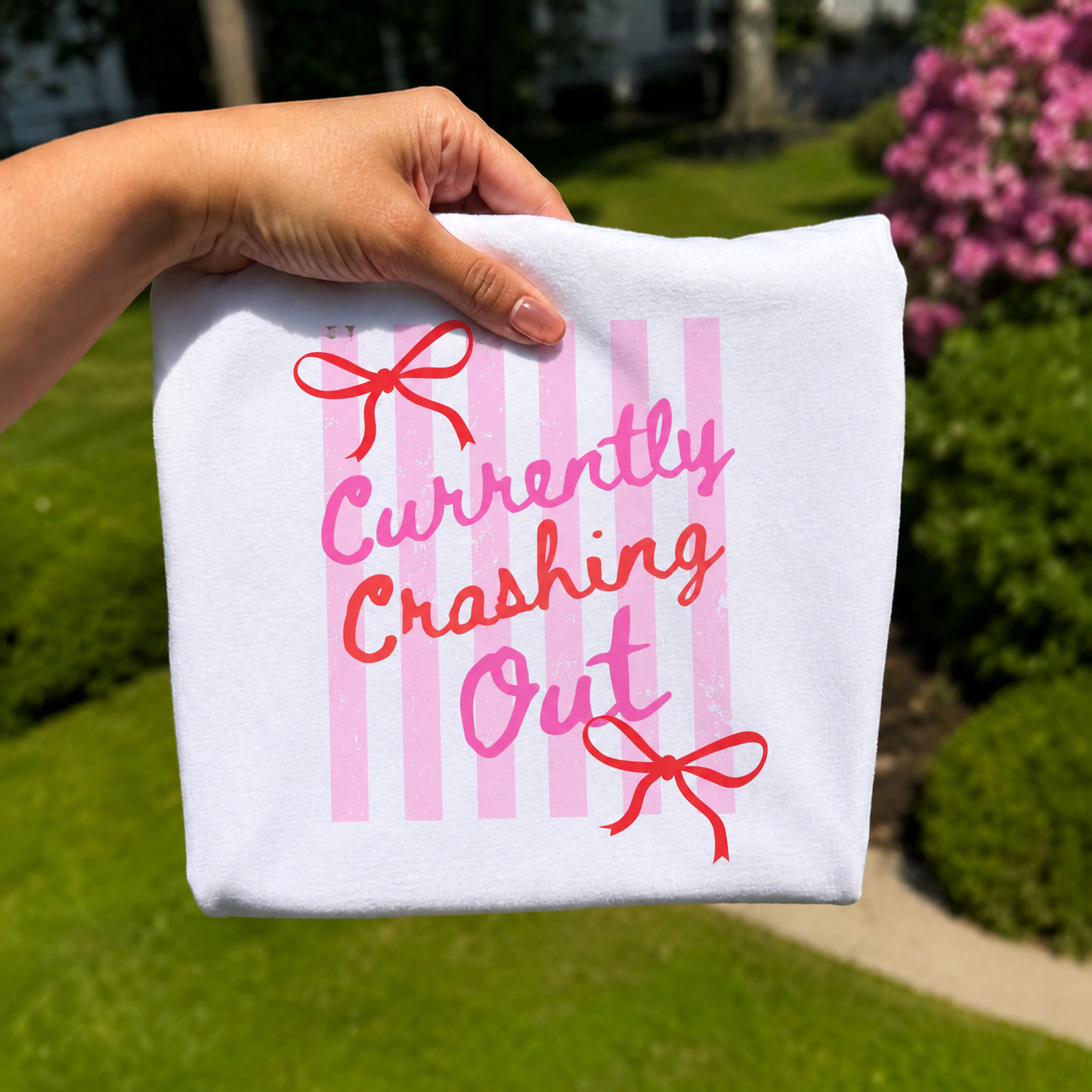 Folded white graphic T-shirt featuring pink striped artwork with the phrase "Currently Crashing Out" accented by bright red bows, photographed outdoors with a hand holding the shirt against a blurred garden background.