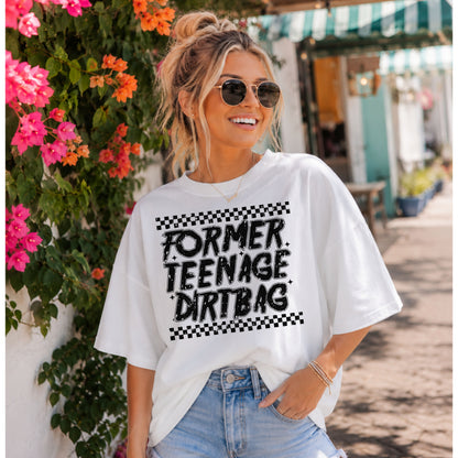 Smiling woman wearing a white oversized graphic T-shirt featuring the phrase "Former Teenage Dirtbag" with distressed black lettering and checkerboard accents, styled with light-wash denim jeans and sunglasses while standing beside colorful bougainvillea on a sunny boutique-lined street.
