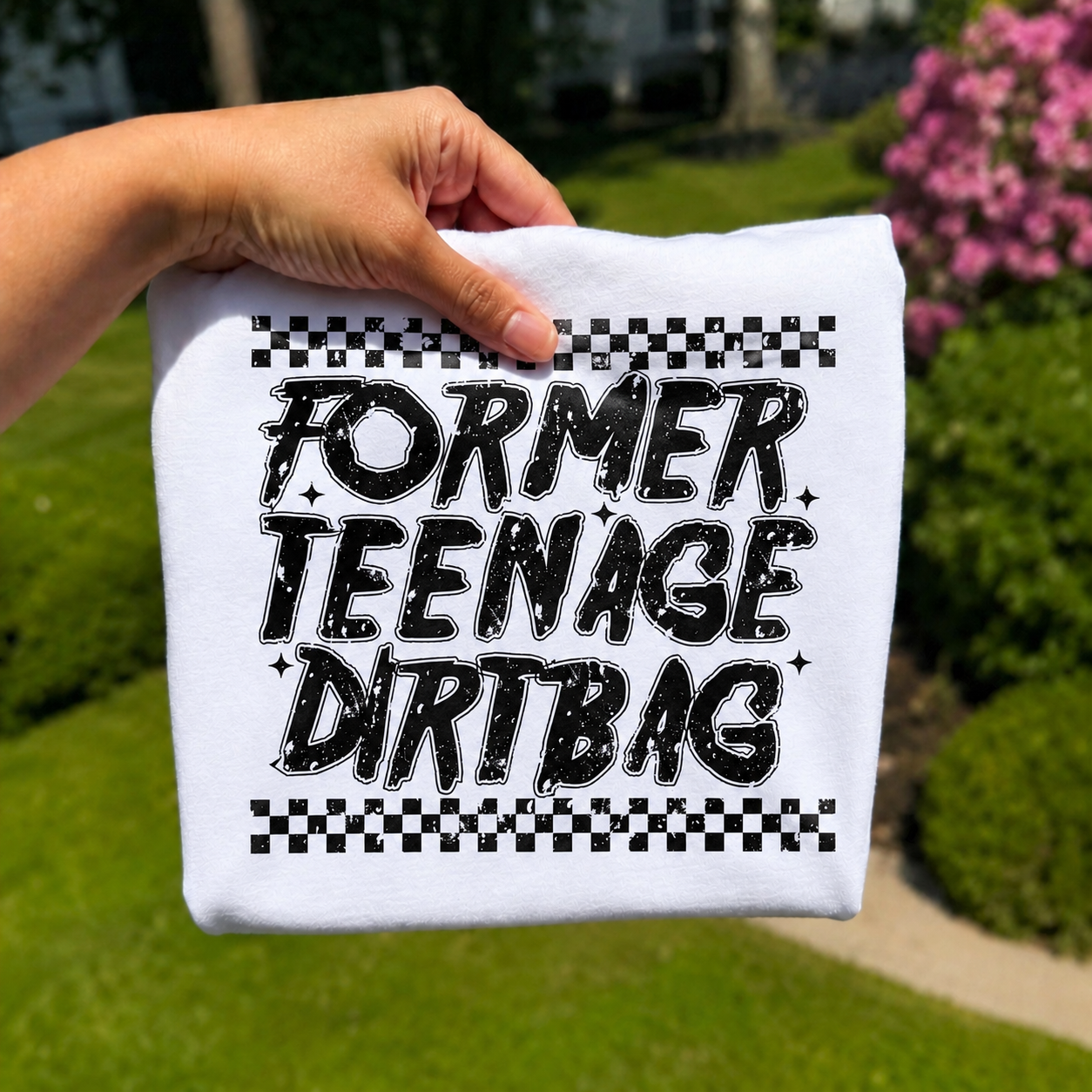 Folded white graphic T-shirt featuring distressed black typography that reads "Former Teenage Dirtbag" with vintage checkerboard accents, photographed outdoors with a hand holding the shirt against a blurred garden background.