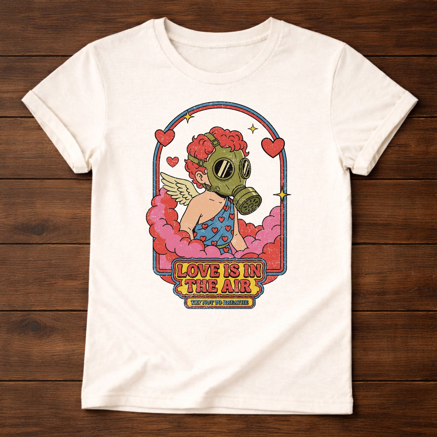 White t-shirt with a graphic design of a person wearing a gas mask and text 'Love is in the Air' on a wooden surface.