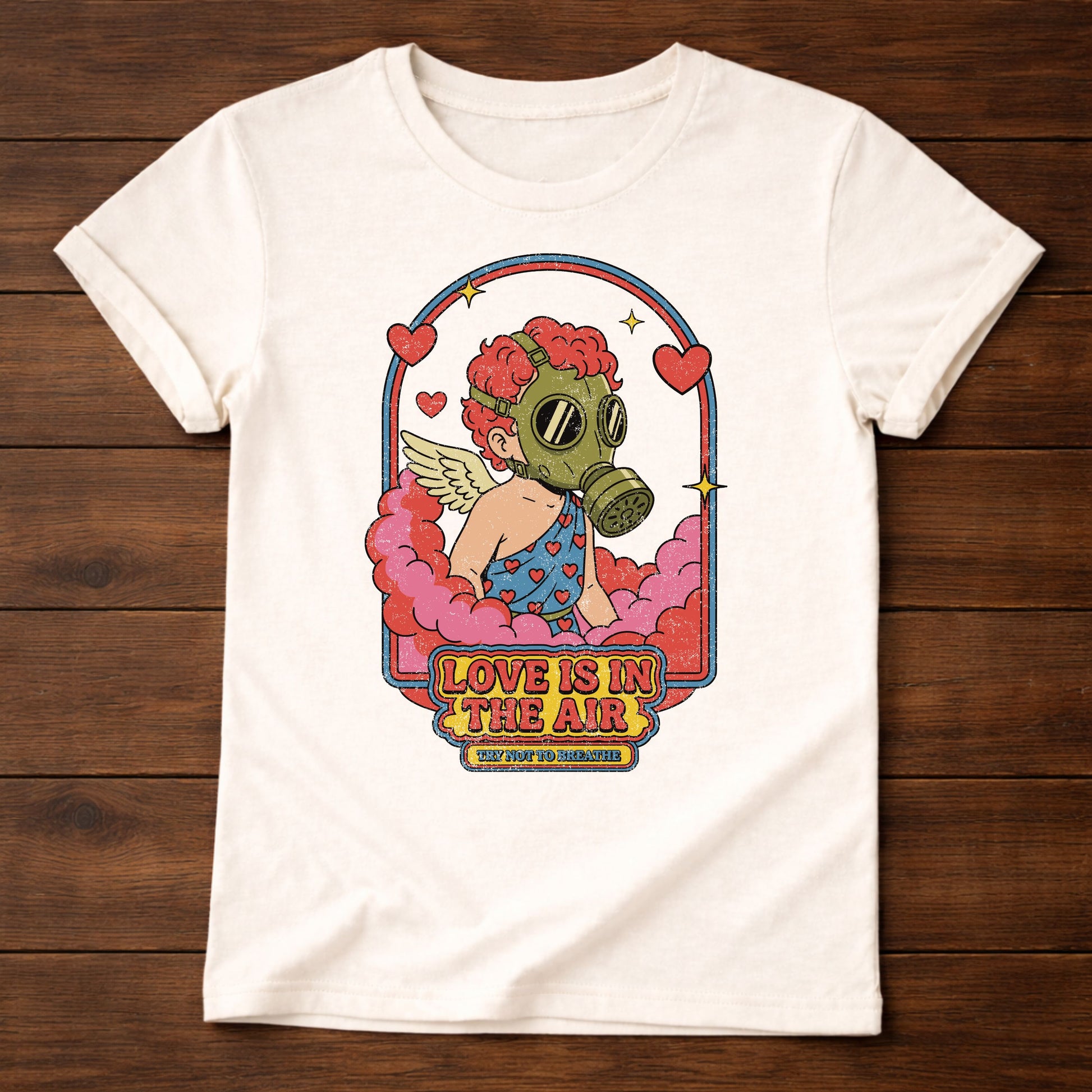 White t-shirt with a graphic design of a person wearing a gas mask and text 'Love is in the Air' on a wooden surface.