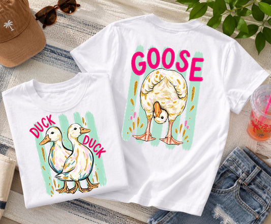 Front-and-back white graphic T-shirt featuring two ducks with the words "Duck Duck" on the front and a goose with the word "Goose" on the back, styled with denim shorts, sunglasses, a beach hat, and a colorful summer drink.