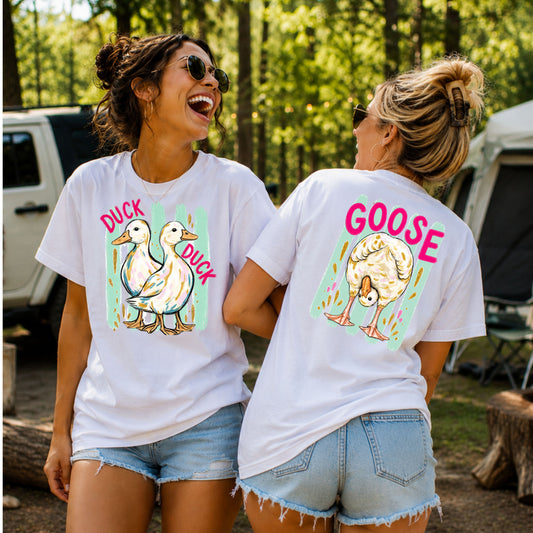 Two women laughing together at an outdoor campground while modeling a white front-and-back graphic T-shirt featuring two ducks with the words "Duck Duck" on the front and a playful goose with the word "Goose" on the back, paired with denim shorts for a fun, casual summer look.