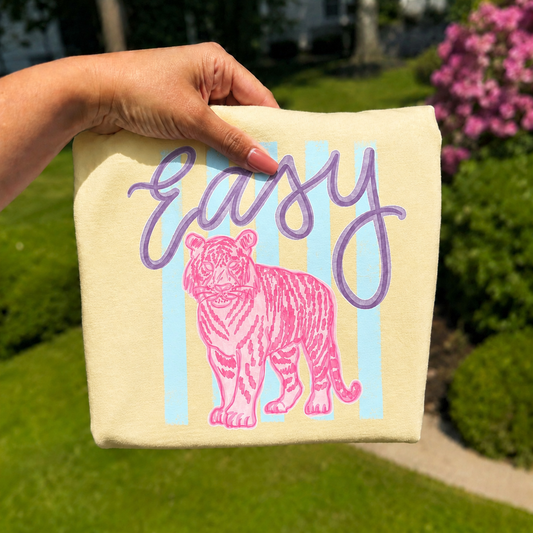Folded butter yellow graphic T-shirt featuring a pink tiger illustration with the phrase "Easy" in lavender script over soft blue pastel stripes, photographed outdoors with a hand holding the shirt against a blurred garden background.