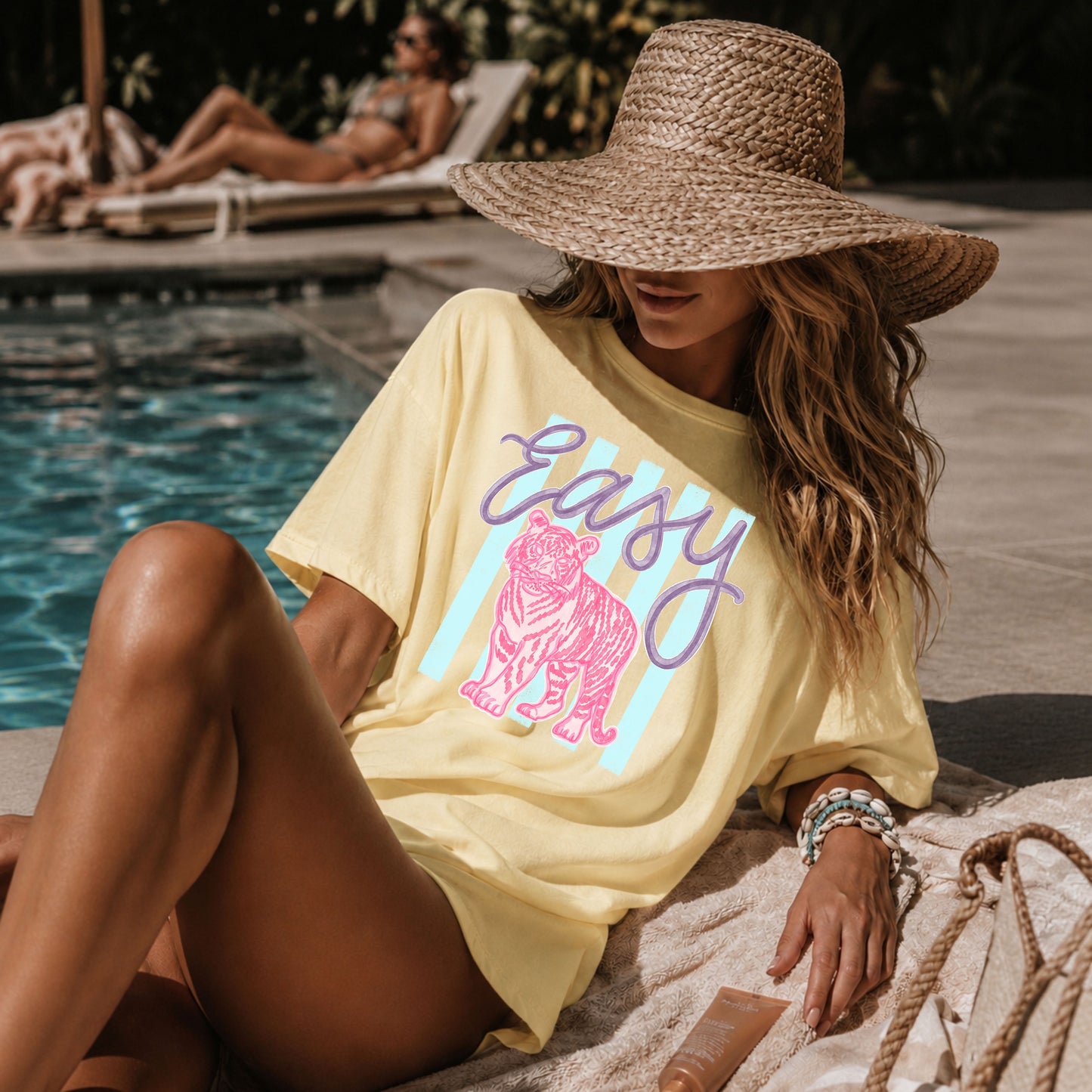 Woman relaxing poolside in an oversized butter yellow graphic T-shirt featuring a pink tiger with the word "Easy" in lavender script over pastel blue stripes, styled with a woven sun hat and beach accessories for a chic coastal summer look.