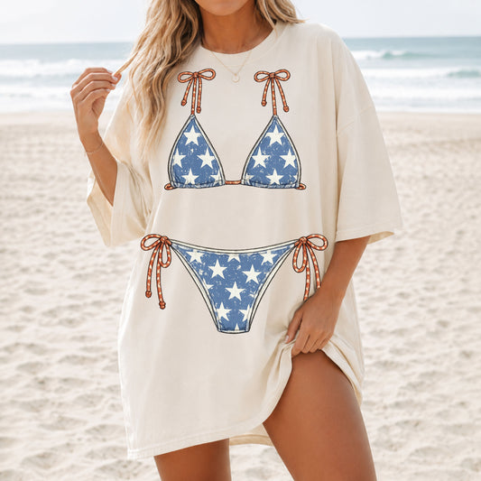 Woman wearing an oversized ivory graphic T-shirt featuring a distressed vintage red, white, and blue bikini illustration while standing on a sunny beach.