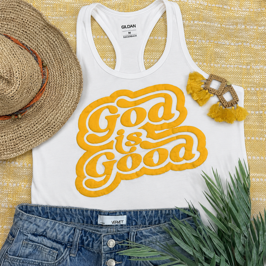 White tank top with 'God is Good' in yellow, paired with denim shorts, a straw hat, and earrings on a textured yellow surface.
