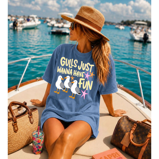 Woman wearing a blue oversized graphic T-shirt featuring the "Gulls Just Wanna Have Fun" design while relaxing on the bow of a boat in a sunny marina, styled with a straw hat, woven tote, and summer accessories.