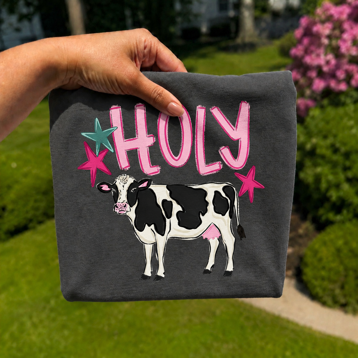 Hand holding a folded charcoal gray graphic T-shirt featuring the phrase "Holy" above an illustrated black and white dairy cow with pink and turquoise star accents, photographed outdoors on a sunny day.