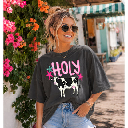 Woman wearing a charcoal gray graphic T-shirt featuring the "Holy Cow" design with a black and white dairy cow, styled with denim shorts and sunglasses while walking along a charming, flower-lined street on a sunny day.