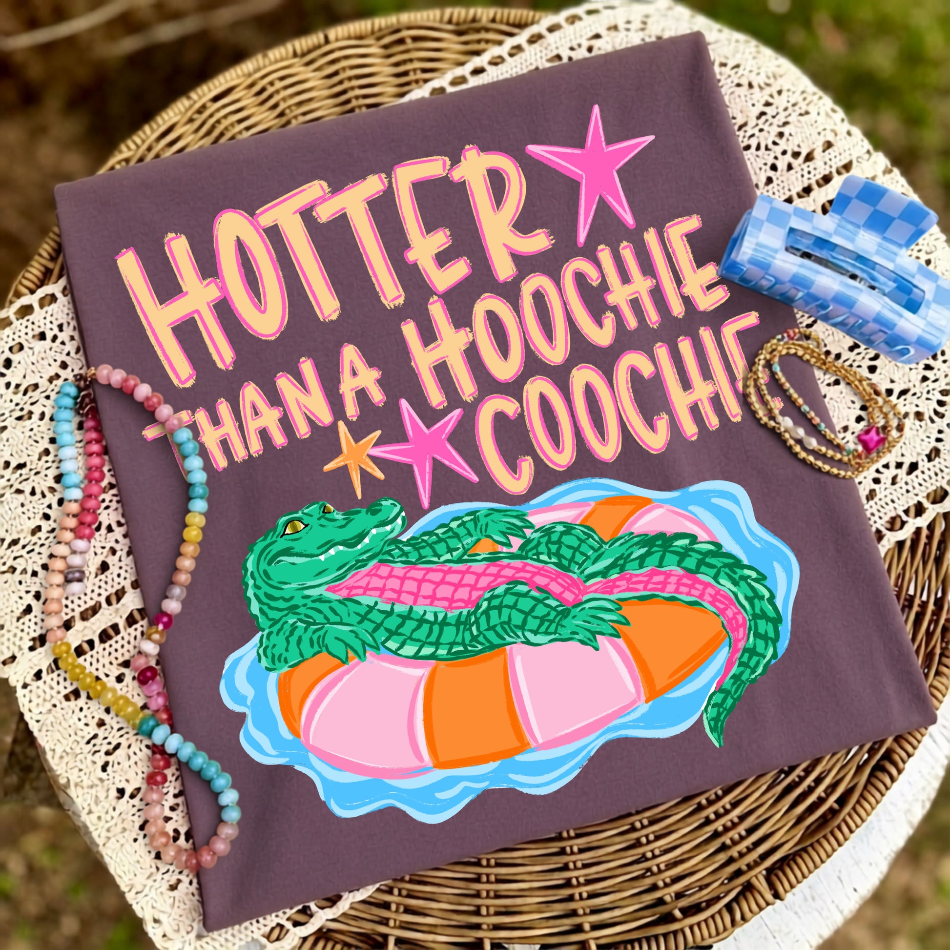 Folded dusk-colored graphic T-shirt featuring the "Hotter Than a Hoochie Coochie" design with a playful alligator floating in a colorful pool tube, styled on a wicker chair with colorful beaded jewelry, a blue hair clip, and a lace throw.
