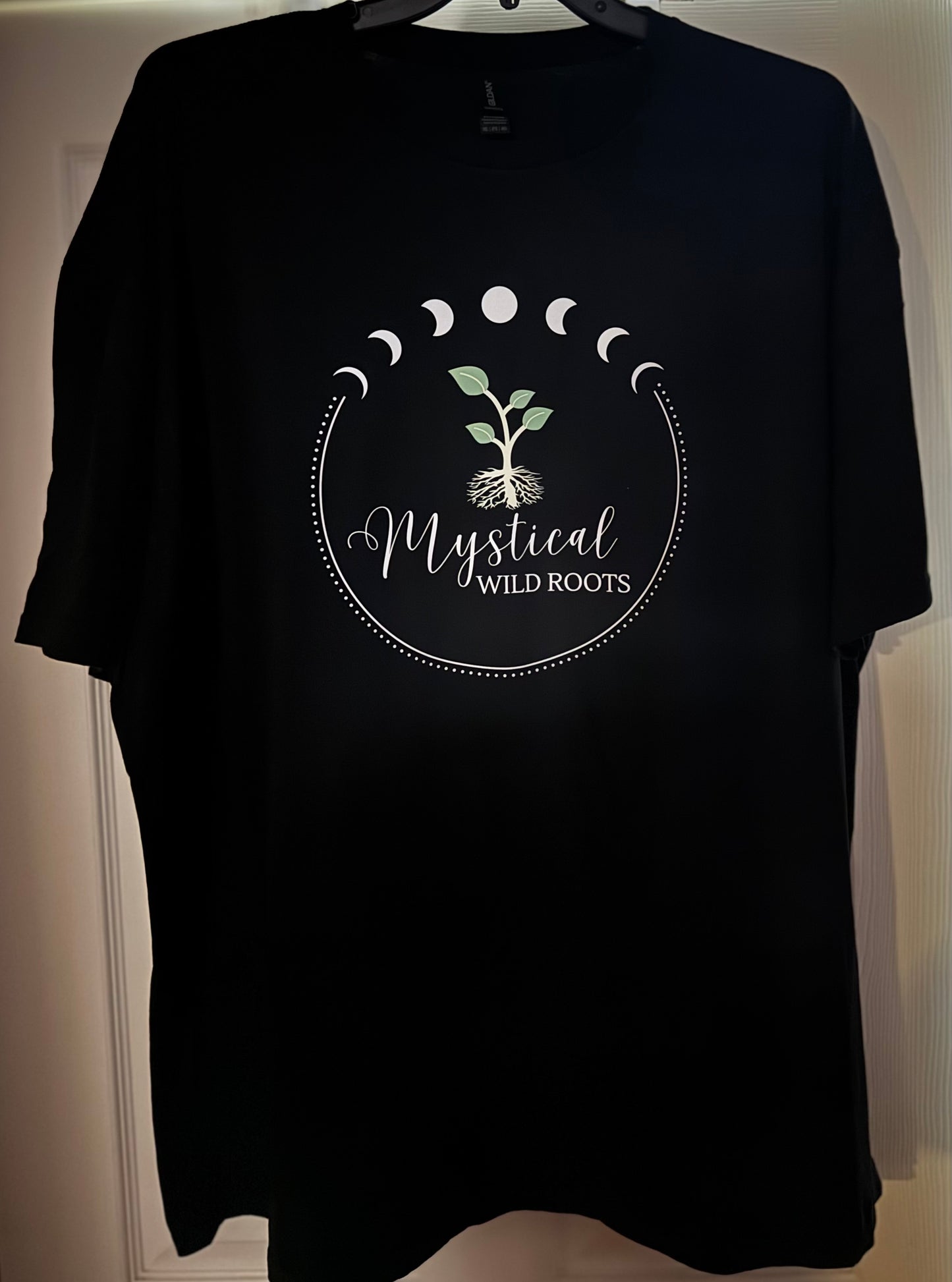 Black t-shirt with 'Mystical Wild Roots' design on a hanger.