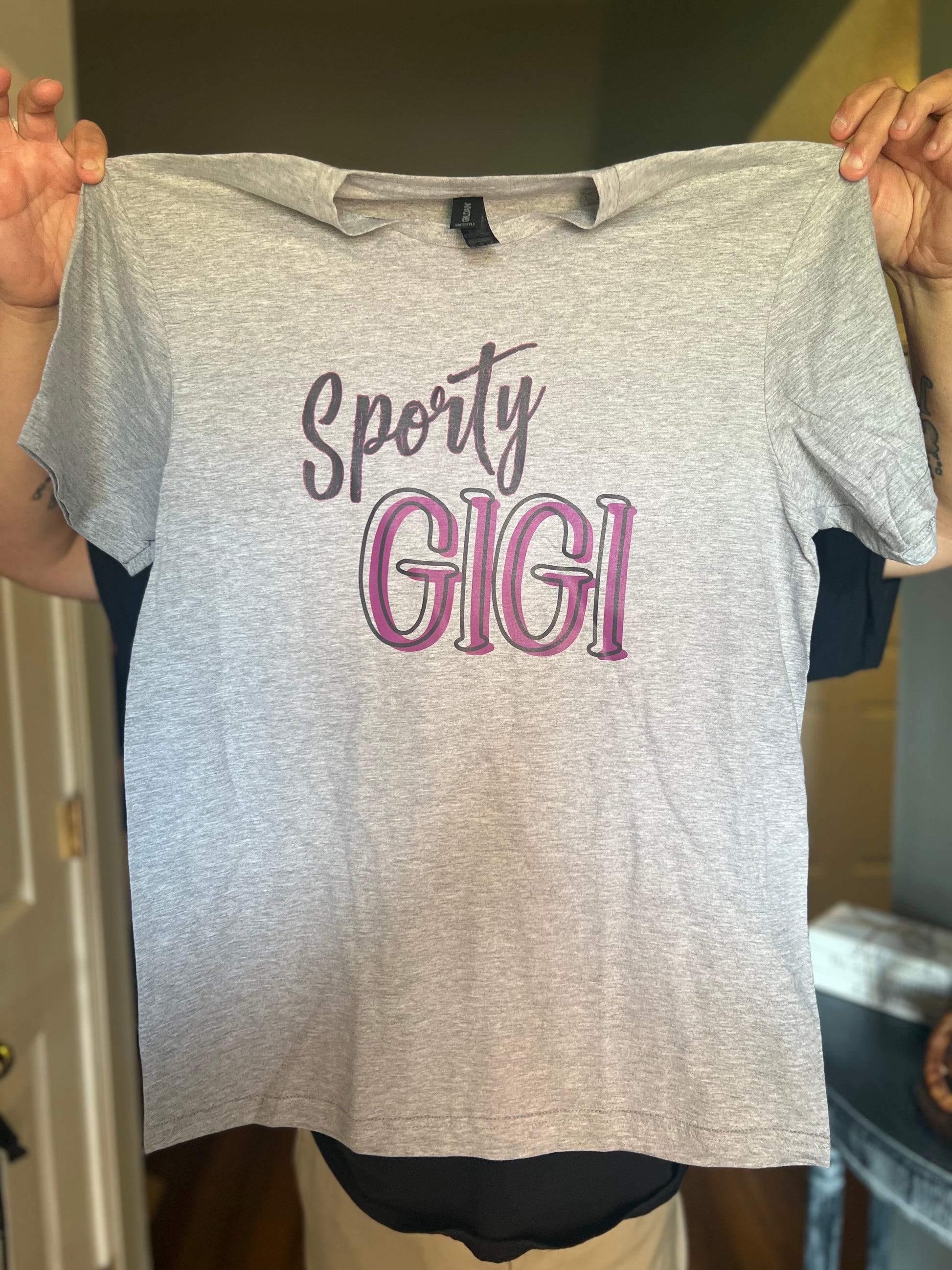 Gray t-shirt with 'Sporty GIGI' text held by a person indoors.