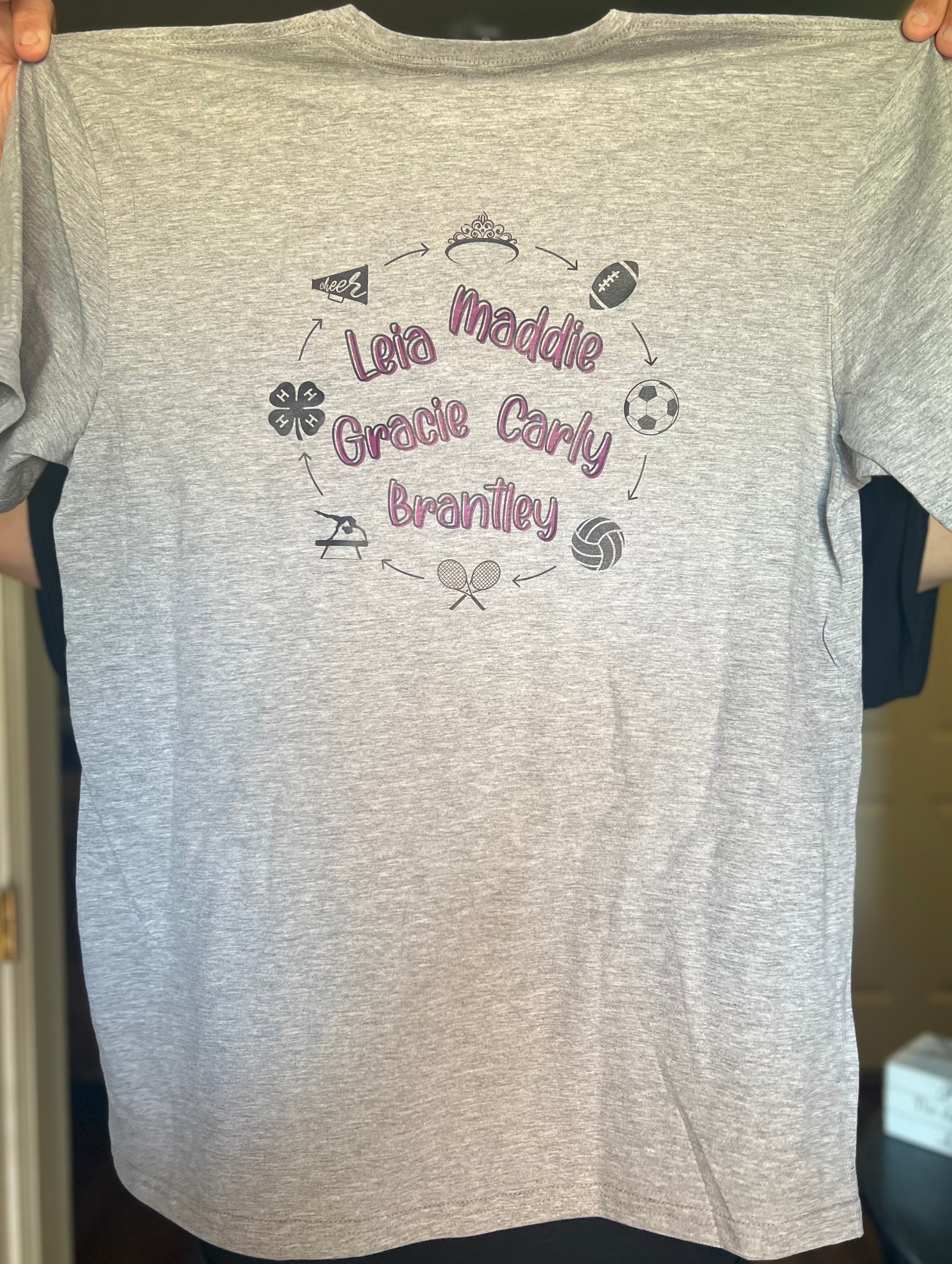 Gray t-shirt with personalized names and sports icons held by a person.