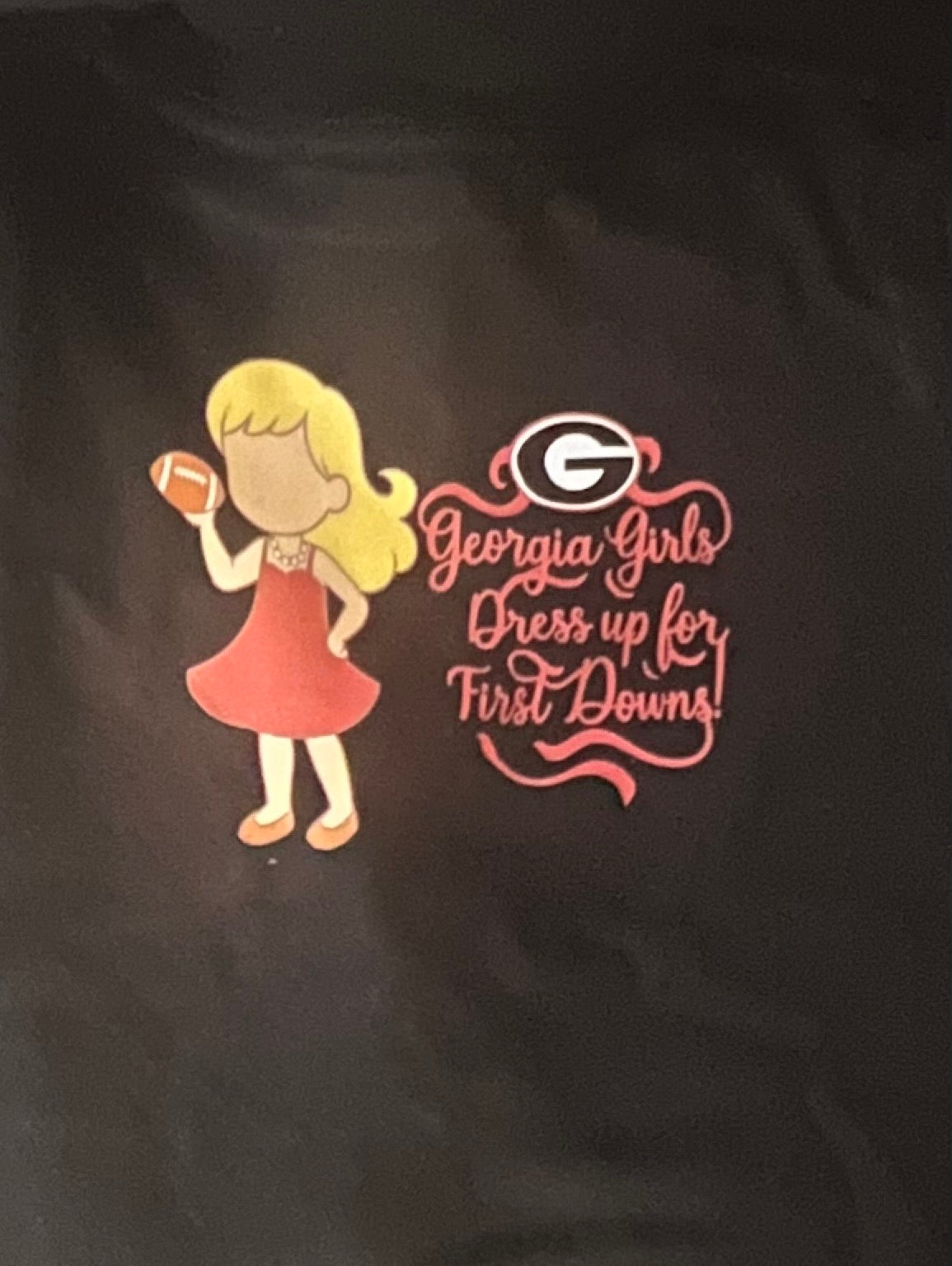 Black shirt with a graphic of a girl in a red dress holding a football and text 'Georgia Girls Dress up for First Downs' on a dark background.