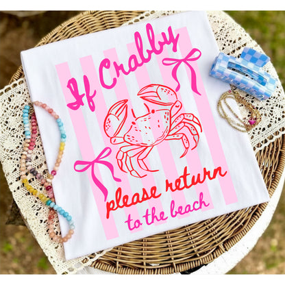 Folded white graphic T-shirt with pink "If Crabby, Please Return to the Beach" crab design displayed on a woven tray with colorful beaded jewelry and beach-inspired accessories.