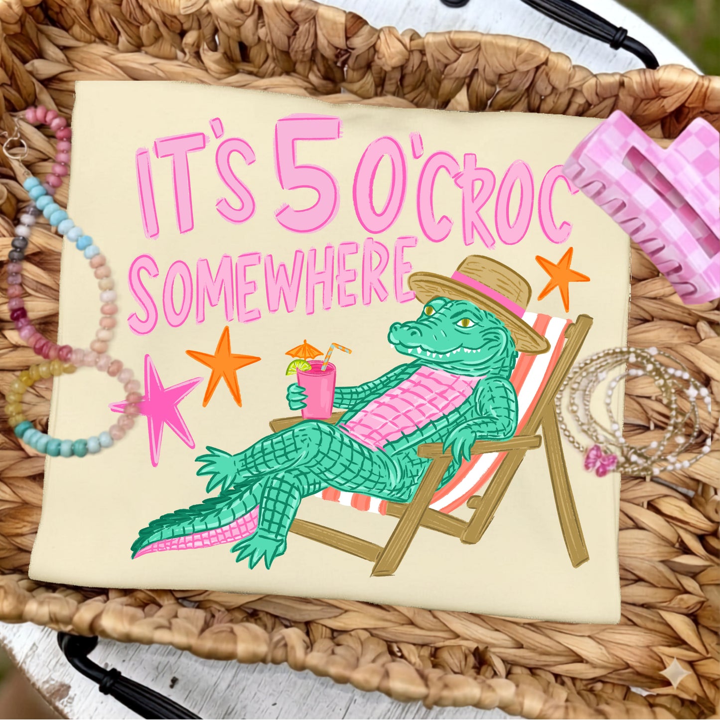 Folded butter yellow graphic T-shirt featuring the colorful "It's 5 O'Croc Somewhere" design with a lounging alligator in a beach chair, styled on a woven tray with pastel beaded bracelets, jewelry, and pink beach-inspired accessories.