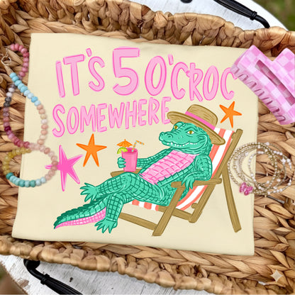 Folded butter yellow graphic T-shirt featuring the colorful "It's 5 O'Croc Somewhere" design with a lounging alligator in a beach chair, styled on a woven tray with pastel beaded bracelets, jewelry, and pink beach-inspired accessories.