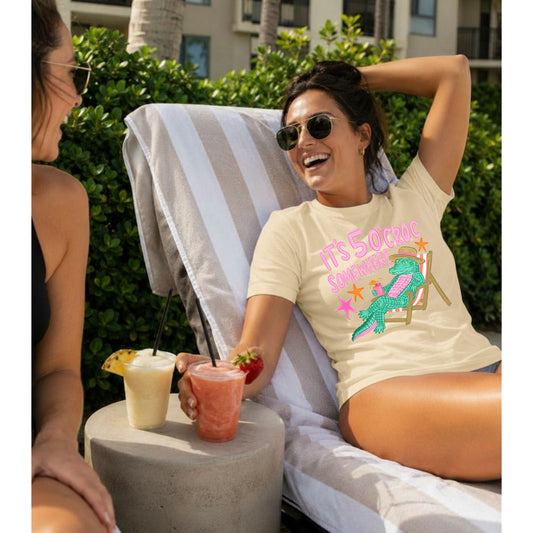 Woman relaxing poolside in a cream graphic T-shirt featuring the colorful "It's 5 O'Croc Somewhere" alligator design while enjoying frozen drinks on a sunny summer afternoon.