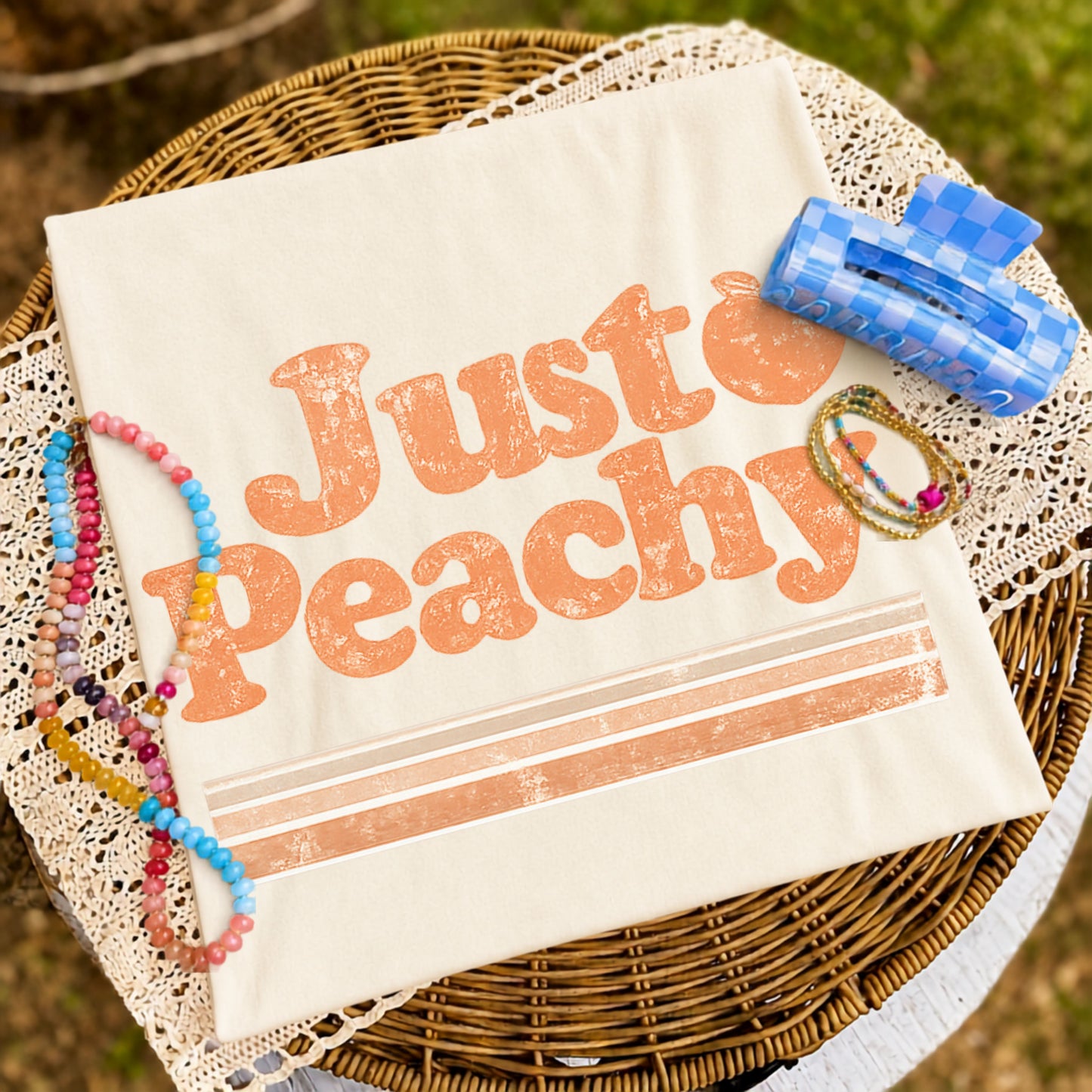 Folded cream graphic T-shirt featuring a distressed "Just Peachy" design displayed on a woven tray with colorful beaded bracelets, delicate jewelry, and a blue checkered hair clip.