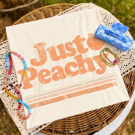 Folded cream graphic T-shirt featuring a distressed "Just Peachy" design displayed on a woven tray with colorful beaded bracelets, delicate jewelry, and a blue checkered hair clip.